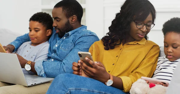 Счастливая африканская американская семья сидит на диване вместе с устройством дома. Мать и отец с двумя маленькими детьми с помощью ноутбука и смартфона . — стоковое фото