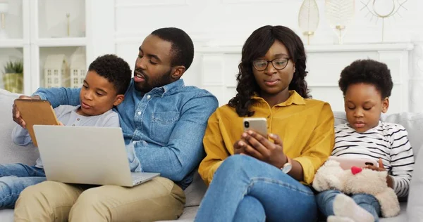 Gelukkige Afrikaans-Amerikaanse familie zit op de bank samen met apparaat thuis. Moeder en vader met twee kleine kinderen met laptop en smartphone. — Stockfoto