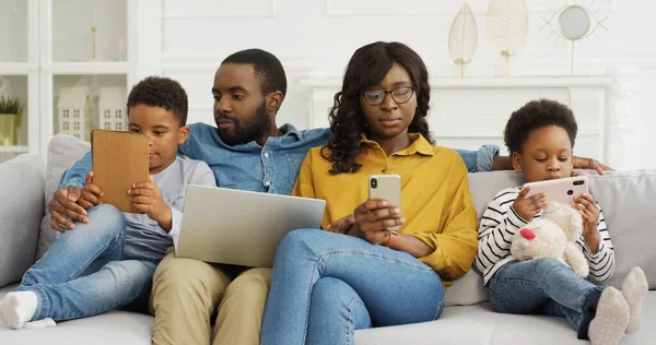 Gelukkige Afrikaans-Amerikaanse familie zit op de bank samen met apparaat thuis. Moeder en vader met twee kleine kinderen met laptop en smartphone. — Stockfoto