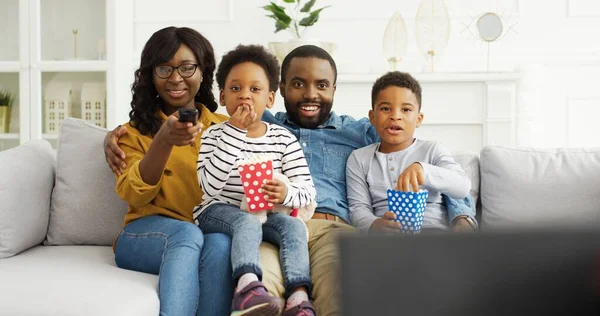 Afro-Amerikaanse ouders met kleine kinderen kijken grappige films of tv-show, eten popcorn, rusten op de bank thuis. — Stockfoto