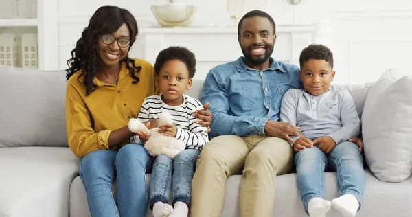Portrét afroamerické rodiny, jak spolu tráví čas doma. Černošky rodiče a malé děti se dívají na fotoaparát sedí spolu na pohovce. — Stock fotografie