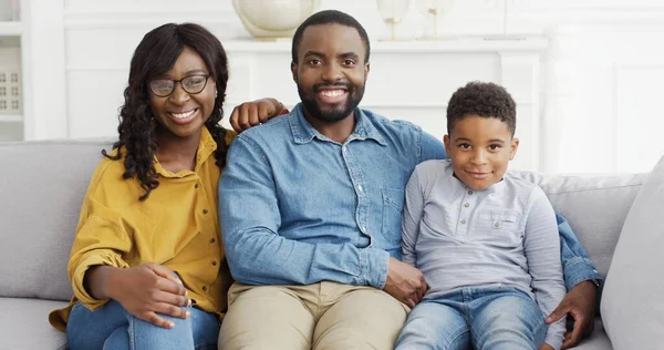 Portret van Afro-Amerikaanse familie die samen tijd doorbrengt thuis. Zwarte ouders en kleine zoon kijken naar de camera zitten samen op de bank. — Stockfoto