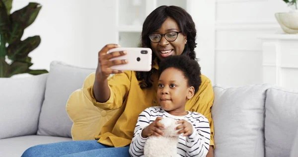 Gelukkig glimlachen Afrikaans amerikaanse moeder en schattig dochtertje nemen selfie op smartphone zitten op de bank in de woonkamer thuis. — Stockfoto