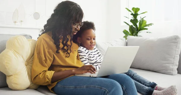 Portret van Afrikaanse Amerikaanse moeder en schattig dochtertje met behulp van laptop zittend op de bank in de woonkamer thuis. — Stockfoto