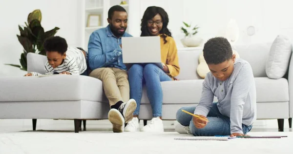 Família afro-americana feliz sentada no sofá junto com o dispositivo em casa. Mãe e pai usando laptop, crianças pequenas irmão e irmã desenho em casa, conceito de tempo de família — Fotografia de Stock