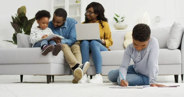 Gelukkige Afrikaans-Amerikaanse familie zit op de bank samen met apparaat thuis. Moeder en vader met laptop, kleine kinderen broer en zus thuis tekenen, familie tijd concept — Stockfoto