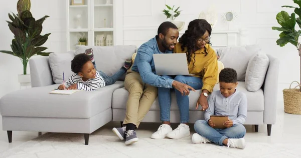 Gelukkige Afrikaans-Amerikaanse familie zit op de bank samen met apparaat thuis. Moeder en vader met laptop, kleine kinderen broer en zus thuis tekenen, familie tijd concept — Stockfoto