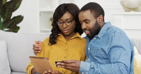 Glad afroamerikansk par man och hustru sitter på soffan med hjälp av surfplatta och kreditkort för online shopping hemma. — Stockfoto