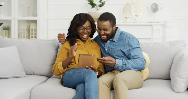 Glad afroamerikansk par man och hustru sitter på soffan med hjälp av surfplatta och kreditkort för online shopping hemma. — Stockfoto