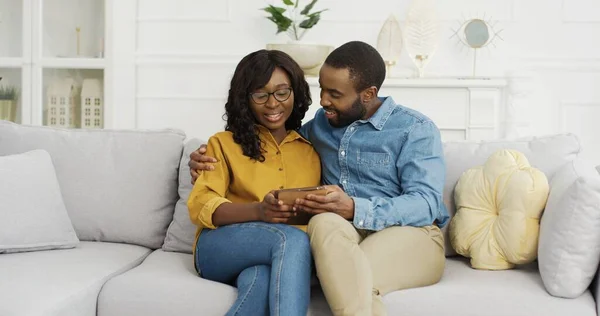 Šťastný africký americký pár manžel a manželka sedí na pohovce pomocí tabletu doma. — Stock fotografie