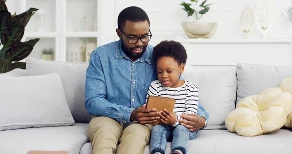 Portret van Afrikaanse Amerikaanse vader zittend met dochter op de bank met behulp van digitale tablet. — Stockfoto