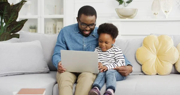 Portret van Afrikaanse Amerikaanse vader zit met dochter op de bank met behulp van laptop kijken video. — Stockfoto