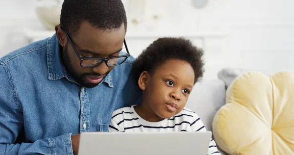 Portret van Afrikaanse Amerikaanse vader zit met dochter op de bank met behulp van laptop kijken video. — Stockfoto