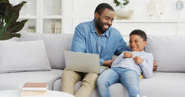 Gelukkig Afrikaans amerikaans man met behulp van laptop met plezier spelen met zoon zitten op bank. — Stockfoto