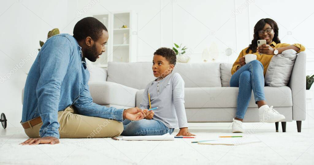 Happy african american family spending time together at home. Cheerful african dad play with little son, young mother using smartphone.