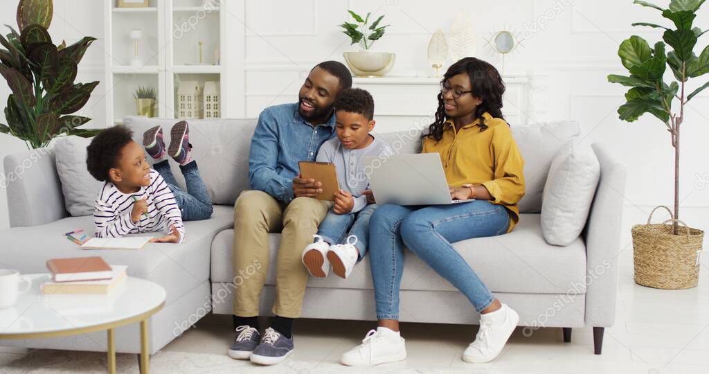 Happy african american family sitting on sofa together with device at home. Mother using laptop, father and son holding digital tablet, cute little girl drawing. Family time concept