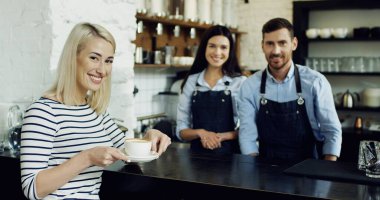 Neşeli genç erkek ve kadın baristler kahve dükkanında çalışıyorlar ve güzel bir kadın elinde latte ile bardaklarını tutuyor. Kahve Dükkanı Konsepti