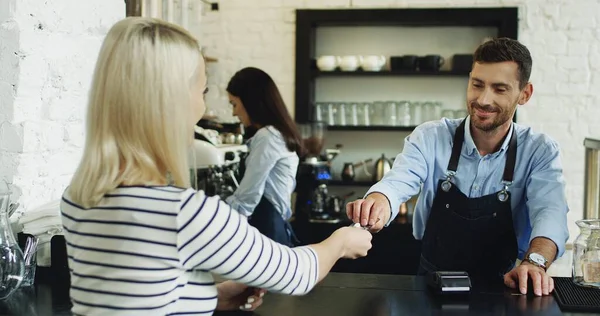 Vista posterior de la mujer rubia pagando con una tarjeta de crédito al camarero en el bar, mientras la camarera sirve su café . —  Fotos de Stock