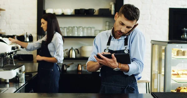 Kaukasische attraktive Kellnerin, die am Telefon spricht und Tablet-Computer an der Bar benutzt, brünette Kellnerin, die hinter ihr Kaffee kocht. — Stockfoto