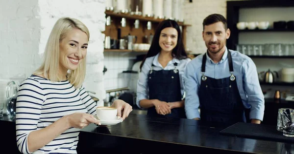 Fröhliche junge männliche und weibliche Baristas arbeiten im Café und die schöne stilvolle Frau hält Tasse mit Latte in der Hand. Café-Konzept — Stockfoto