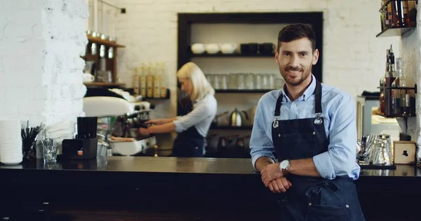 Portraitaufnahme des gutaussehenden lächelnden Kellners, der an der Bar steht und lehnt, während die blonde attraktive Kellnerin hinter ihm Kaffee zubereitet. — Stockfoto