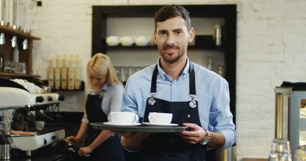 Portraitaufnahme des gutaussehenden kaukasischen Barmanns, der Tassen Kaffee hält und in die Bar geht, während seine Mitarbeiterin hinter der Kaffeemaschine mehr zubereitet. — Stockfoto