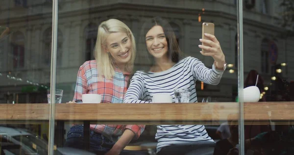 Hermosas dos mujeres posando y tomando buenas fotos selfie en la cámara del teléfono inteligente mientras se sienta con café en la cafetería . — Foto de Stock
