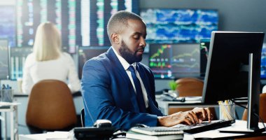 Borsada çalışan Afro-Amerikan borsa simsarı iş ekibinin arka planındaki bilgisayarı kullanıyor ve veri, saat numaraları ve grafikleri gösteren birden fazla monitör kullanıyor..