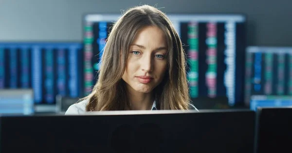 Close up portrait of female trader or broker working at stock exchange office using computer on background of multiple monitors showing data, ticker numbers and graphs.
