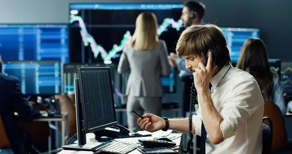 在股票交易所办公室工作的男性股票交易员 他的商业团队背景下的会说话的电话 以及显示数据 打卡号和图表的多个监视器 — 图库照片