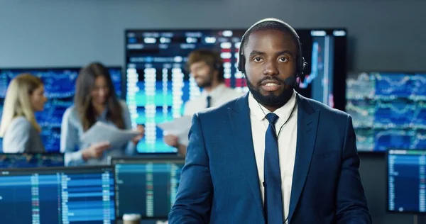 Portrait of african american trader or brokers working at stock exchange office using headset and looking at camera on background of her business team.