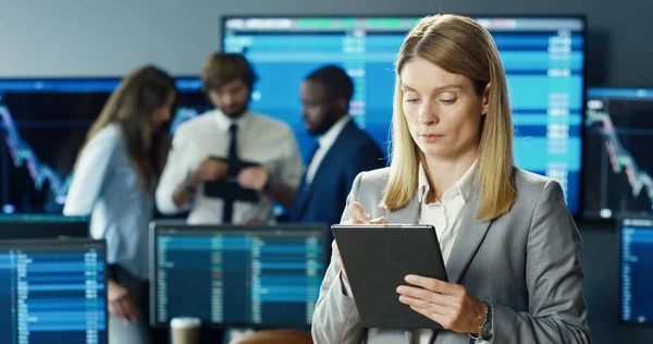在股票交易所办公室工作的女性股票交易员使用数字平板电脑 背景上有多个显示器 显示数据 打卡号和图表 — 图库照片