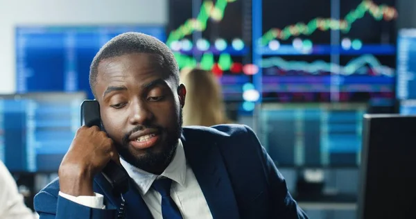 Portrait of african american trader or broker working at stock exchange office and talking on phone background of multiple monitors showing data, ticker numbers and graphs.