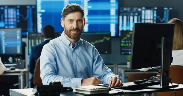 在股票交易所工作的男性股票交易员 在他的商业团队的背景下看着摄像机 还有显示数据 打卡号和图表的多个监视器 — 图库照片