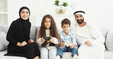 Küçük çocukları olan neşeli Arap ebeveynler kanepede oturup, joysticklerle video oyunu oynuyorlar. Anne ve babasıyla sevimli Arap çocukları evde kontrolörlerle video oyunları oynuyorlar..