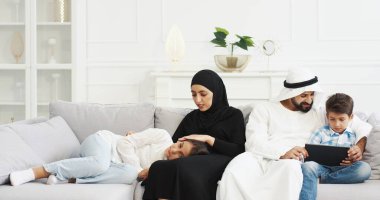 Çocuklu bir Arap ailesi oturma odasında birlikte vakit geçiriyor. Tesettürlü müslüman bir anne, küçük kızını okşuyor. Başı annelerin dizlerinin üzerinde yatıyor. Küçük oğlu olan Arap bir baba tablet kullanıyor ve oynuyor..