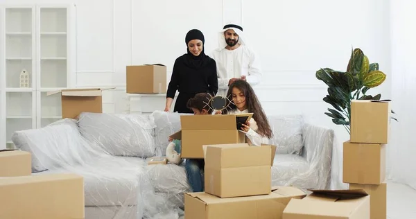 Muslim Arabic parents in traditional clothes in living room with small cute kids unpacking carton boxes. Arabic family with children moving in new house after repairment. Replacing in new apartment. — Stock Photo, Image