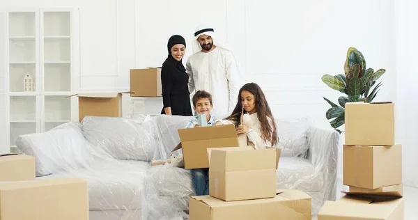 Ευτυχισμένοι Άραβες γονείς φέρνουν πράγματα στο δωμάτιο και μικρά χαριτωμένα παιδιά ξεπακετάρουν χαρτοκιβώτια. Οικογένεια με παιδιά που μετακομίζουν σε νέο σπίτι μετά την επισκευή. Αγόρι και κορίτσι με μητέρα και πατέρα αλλάζουν σπίτι. — Φωτογραφία Αρχείου