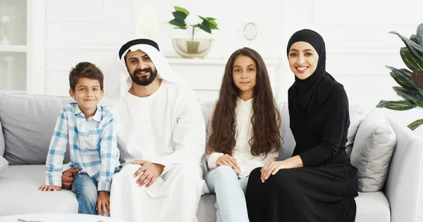 Porträt einer fröhlichen muslimischen Familie mit zwei Kindern, die auf der Couch sitzen und freudig in die Kamera lächeln. Kleine süße Jungen und Mädchen mit Mutter und Vater zu Hause. Arabische Eltern mit kleinen Kindern umarmen sich. — Stockfoto