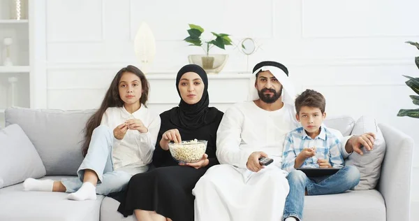 Pais árabes alegres com crianças pequenas sentadas no sofá, sorrindo, assistindo filme na TV e comendo pipocas. Bonito árabe crianças pequenas com mãe e pai passar o tempo juntos em casa . — Fotografia de Stock