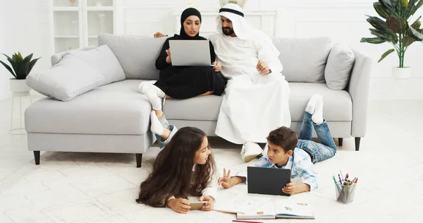 Arabskie szczęśliwe dzieciaki leżące na podłodze i bawiące się gadżetami. Mała siostra i brat za pomocą smartfona i tabletu. Para rodziców siedzi na kanapie z laptopem. Arabowie matka i ojciec odpoczywają. — Zdjęcie stockowe