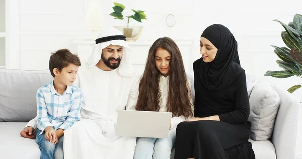 Мусульманские счастливые маленькие дети сидят с родителями на диване и смотрят видео на ноутбуке. Мать-отец с маленькими детьми, дочь и сын разговаривают, используя устройство. Арабская семья в изоляции. — стоковое фото