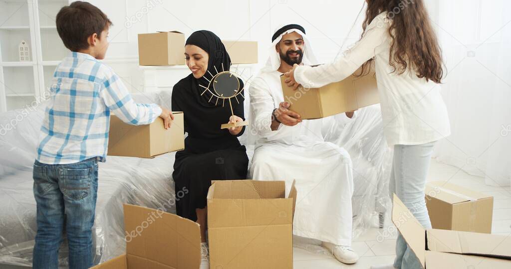 Arabian married couple in traditional Middle Eastern clothes siting on couch and unpacking carton houses. Kids helping to parents when family moving in new accomodation. Arabs in traditional clothes.