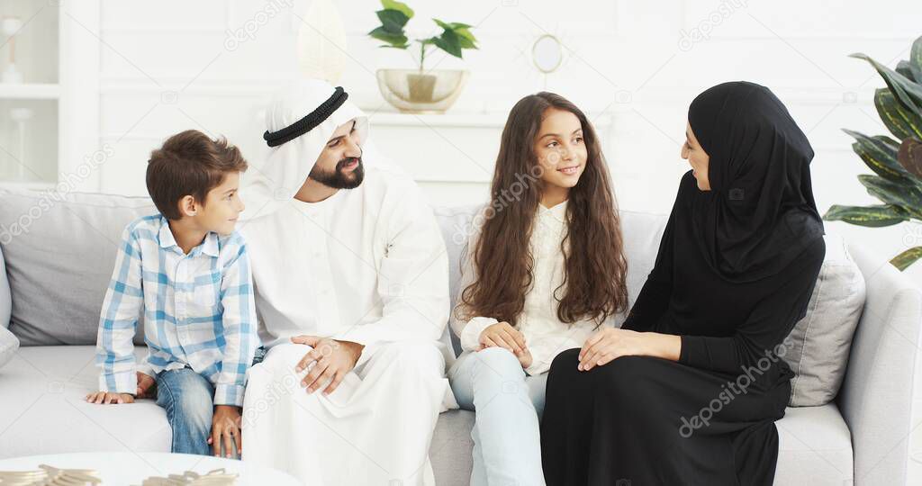 Portrait of cheeful Muslim family with two kids sitting on couch and smiling joyfully to camera. Small cute boy and girl with mother and father at home. Arabian parents with little children hugging.