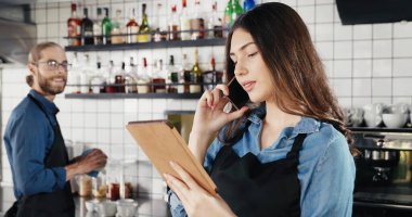 Güzel beyaz kadın barmen telefonda konuşuyor ve barda tablet cihazını tutuyor. Adam arka planda çalışıyor ve kıza gülümsüyor. Garson kız cep telefonuyla konuşuyor.