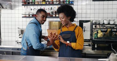 Önlüklü güzel Afro-Amerikalı bayan garson tezgahta duruyor, tablet cihazı kullanıyor ve erkek garsonla konuşuyor. Barmen 'e barmen gösterirken kadın barfiks tıkırdıyor..
