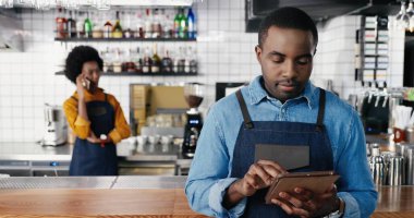 Kafede tablet kullanan ve kameraya gülümseyen Afrikalı Amerikalı erkek barmenlerin portresi. Yakışıklı garson. Cihazla internete bağlanıp kayıyor. Garson arka planda cep telefonuyla konuşuyor..