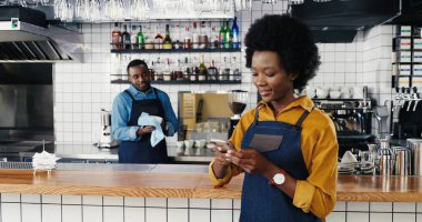 Güzel Afro-Amerikalı garson barda cep telefonunu dinleyip kaydırıyor. Barikat kuran kadın akıllı telefondan mesaj atıyor. Garson arka planda bardakları siliyor. Kafede temizlik bardakları.