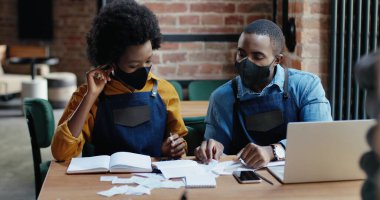 Maskeli Afro-Amerikalı çift restoran hesaplamaları yapıyor ve faturaları kontrol ediyor. Harcamalar ve hasarlar hesaplanıyor. Tecritten sonra tekrar açın. Dizüstü bilgisayarda sayılıyor Küçük girişimcilik