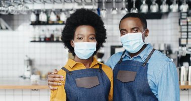 Siyahi Amerikalı neşeli barmenlerin portresi. Maskeli iki barmen birbirlerine sarılıyorlar, kameraya mutlu bir şekilde gülümsüyorlar. Kucaklaşan kafede mutlu garson ve garson. Barristas kadınını kucaklayan neşeli bir adam